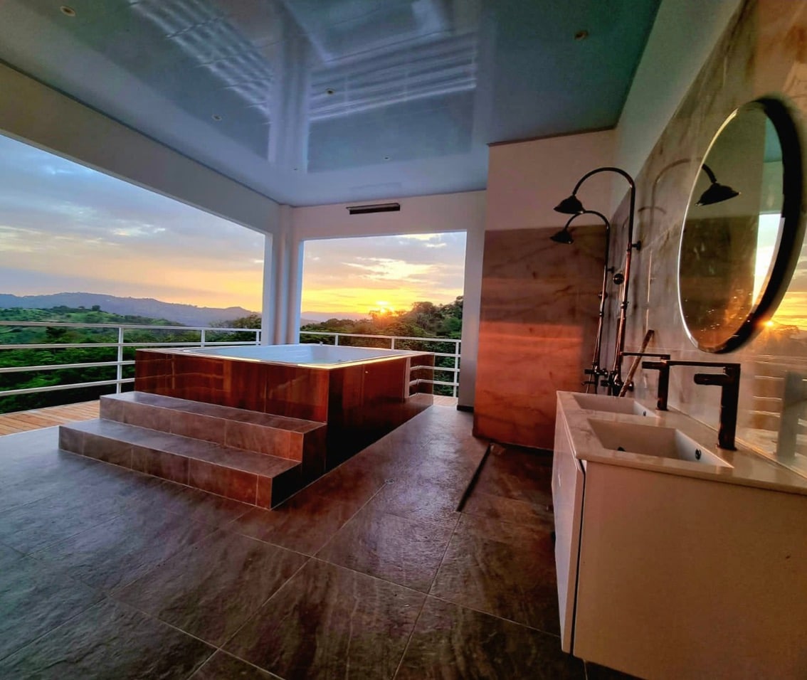 A spacious bathroom features a large private Jacuzzi with steps leading up to it, set against a backdrop of sunset-lit hills. A modern sink and faucet complement the elegant design, while expansive windows frame the scenic view.