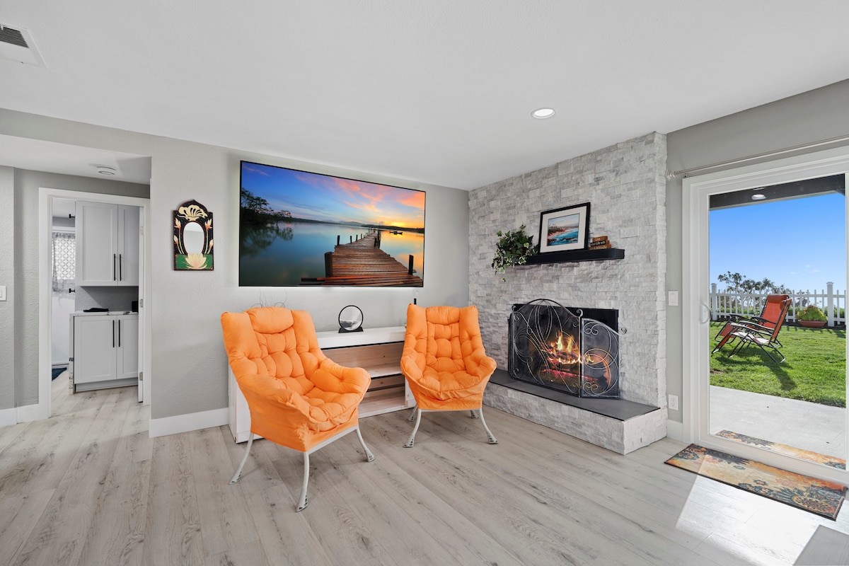 A bright living area is equipped with two orange, tufted chairs facing a sleek fireplace. A large wall-mounted television displays a scenic view. Natural light fills the space from the nearby sliding glass doors leading to a private outdoor area.