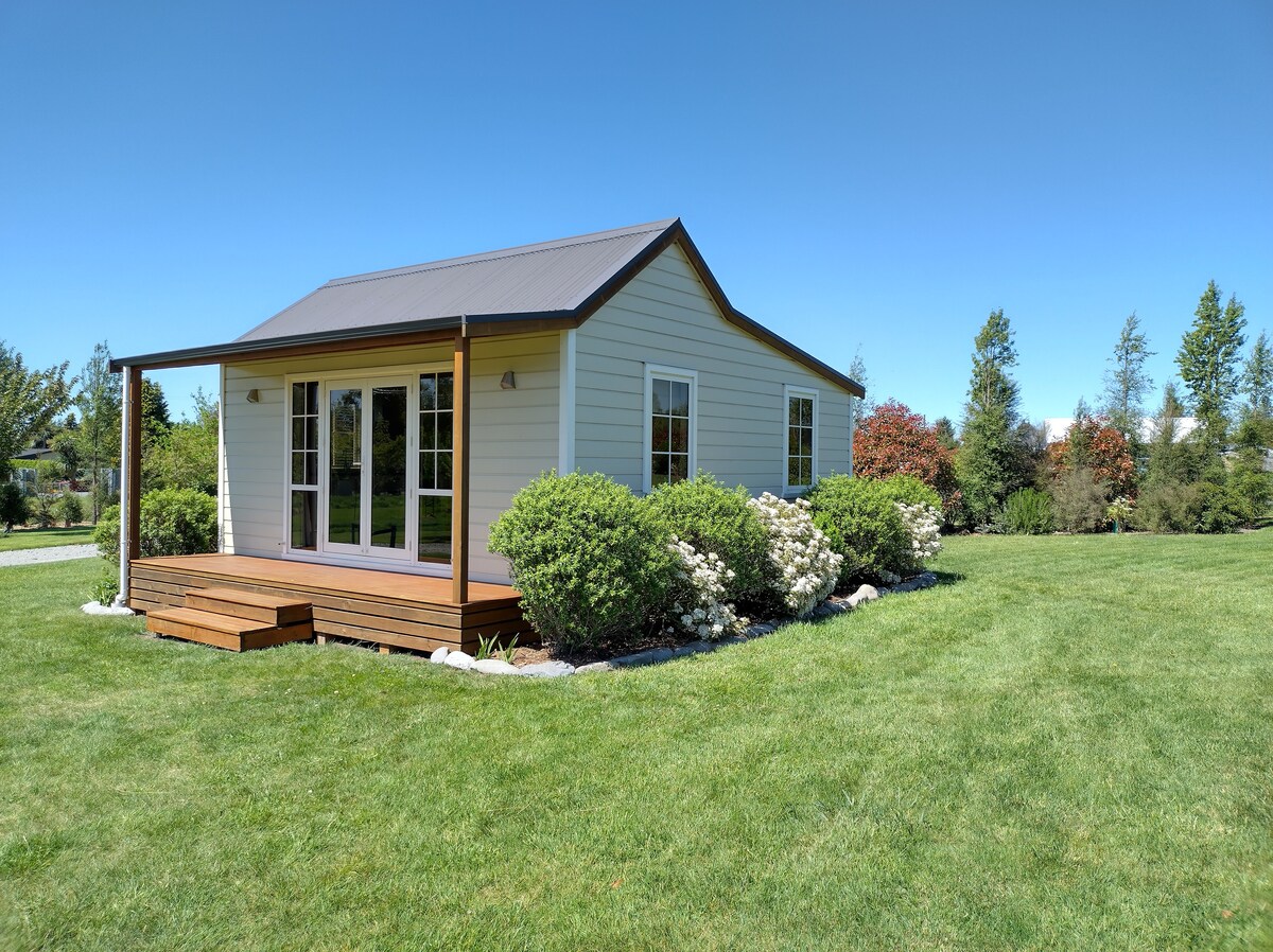 A well-maintained cottage is set on a lush green lawn, framed by neatly trimmed shrubs and flowering bushes. Large glass doors provide a view into the inviting interior, while a wooden deck extends from the front, offering a space to relax outdoors.