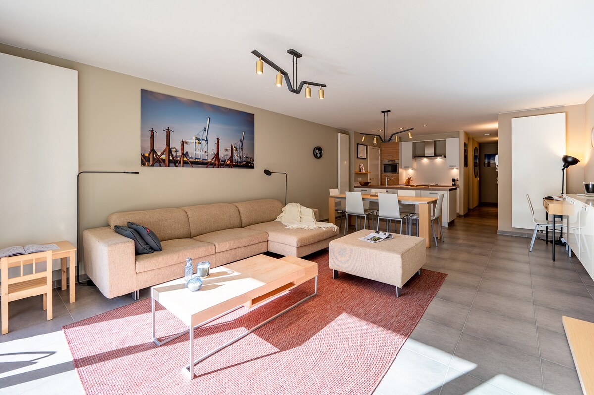 A spacious living area is filled with natural light, featuring a large sectional sofa and coffee table on a textured rug. An open dining area with modern chairs is visible, along with a well-equipped kitchen in the background, complemented by artistic wall decor.