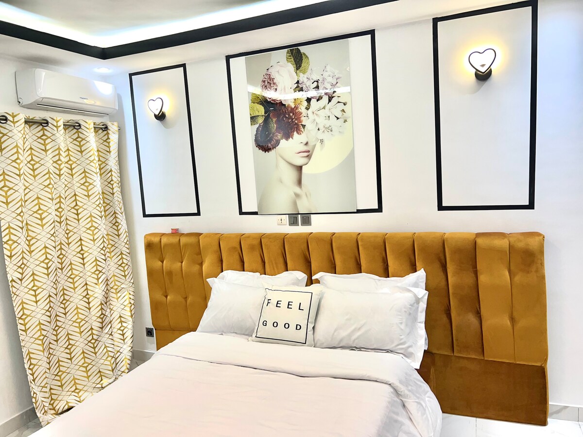 A serene bedroom features a plush, upholstered headboard in warm tones. A soft bed with white linens is complemented by a decorative pillow that reads 'FEEL GOOD.' Artwork depicting a figure adorned with flowers is displayed above the headboard, while patterned curtains add texture to the space.