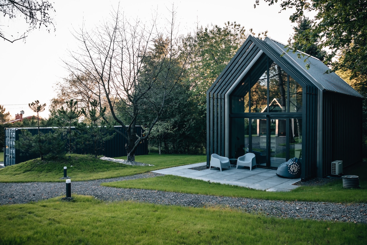 A modern two-level house features sleek lines and large glass panels, allowing for abundant natural light. Outdoor seating is provided on a paved terrace, surrounded by lush greenery and mature trees. A pathway leads into the inviting garden area.