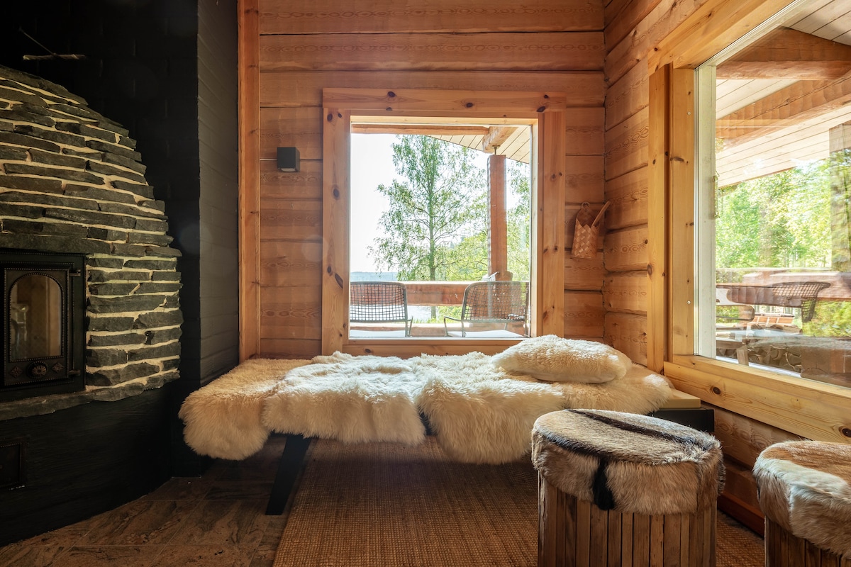 A cozy corner features a comfortable lounging area with a soft fur throw over a minimalist bed frame. Natural light filters through large windows, offering views of the surrounding trees and outdoor seating. A sleek stone fireplace adds warmth to the wooden interior.