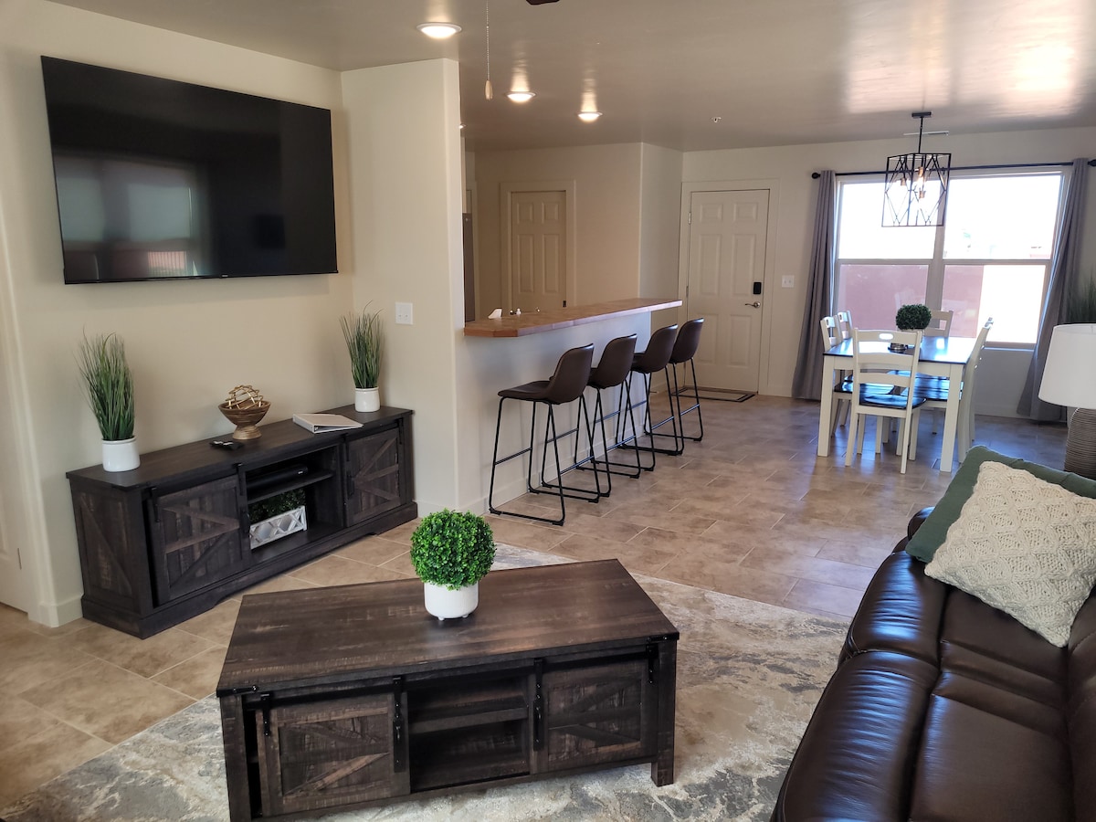 The living area is furnished with a dark coffee table and a spacious sectional couch. A large flat-screen TV is mounted on the wall. A bar with tall stools is visible in the background, along with a dining table set for four. Natural light filters through the windows.
