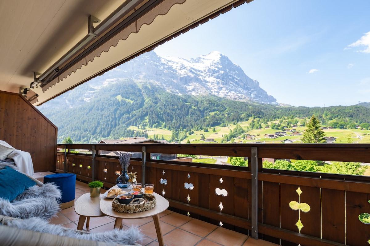 A spacious balcony is presented with views of the Eiger mountain and lush green hills. A wooden table is set with drinks and light snacks, surrounded by various seating options including cushions. The clear blue sky adds to the serene atmosphere of the outdoor space.