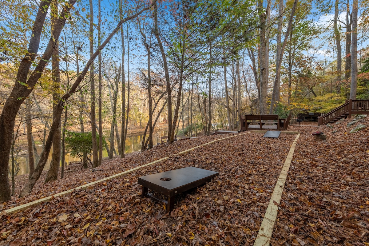 A serene outdoor area features two cornhole boards placed on a carpet of fallen leaves, surrounded by trees displaying autumn foliage. The calm river view is visible in the background, providing a natural setting for relaxation and entertainment.