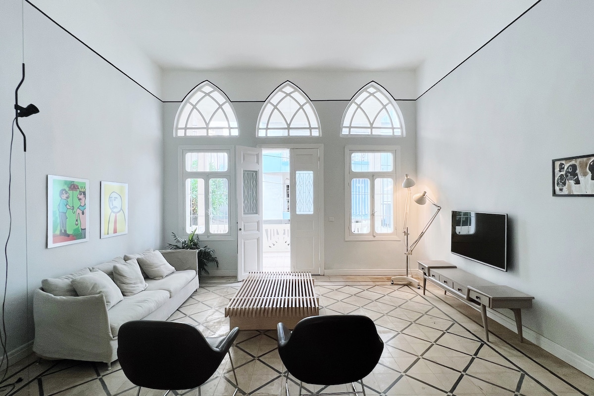 A spacious living room features large arched windows allowing natural light to fill the space. A neutral-colored sofa is paired with two modern black chairs, while a minimalist coffee table sits at the center. Decorative art pieces adorn the walls, contributing to the room's charm.