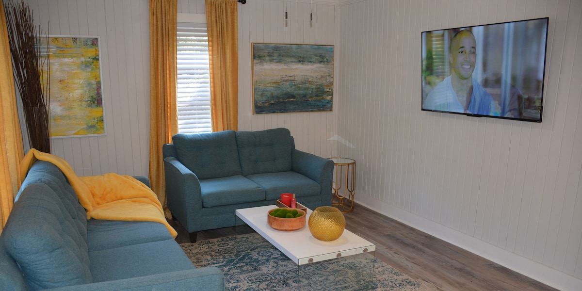 A modern living room is presented, featuring a light blue sofa and a matching loveseat. A white coffee table holds decorative items, including a gold bowl and a small plant. Colorful artwork adorns the walls, and a large television is mounted across from the seating area.