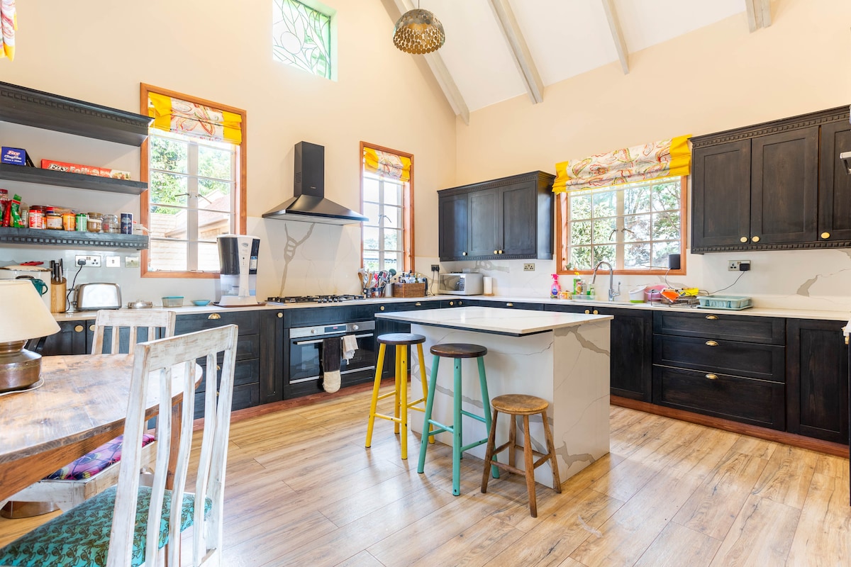A modern kitchen featuring dark cabinetry and a central island. Colorful bar stools, including yellow, complement the space. Natural light floods through large windows adorned with bright curtains, while the wooden floor adds warmth to the open layout.