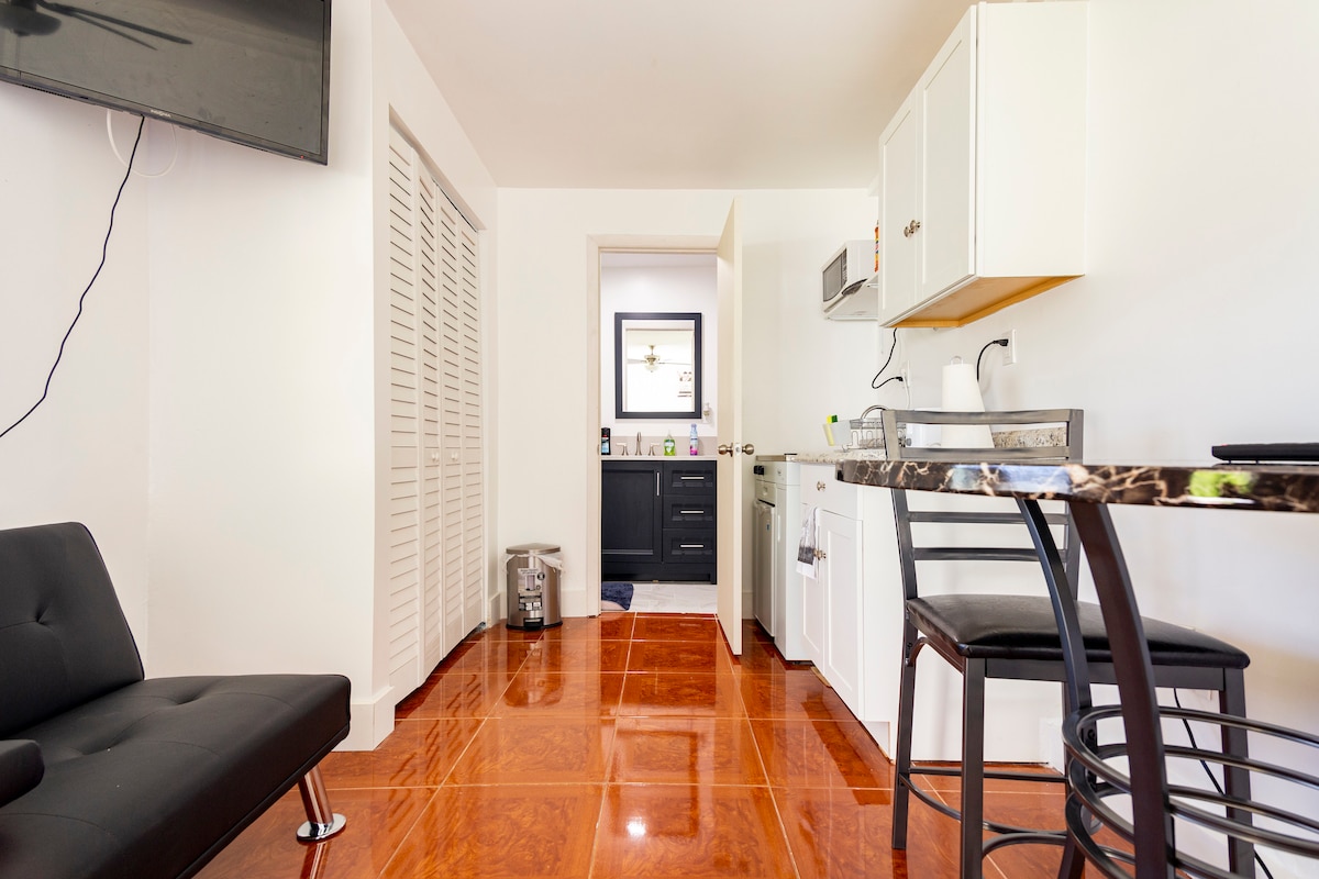 An open living space features glossy tiled flooring and a modern kitchenette, equipped with essential appliances. A dining table with stylish chairs sits beside a sleek black couch. Nearby, a door leads to a bathroom, enhancing the functional layout of the area.