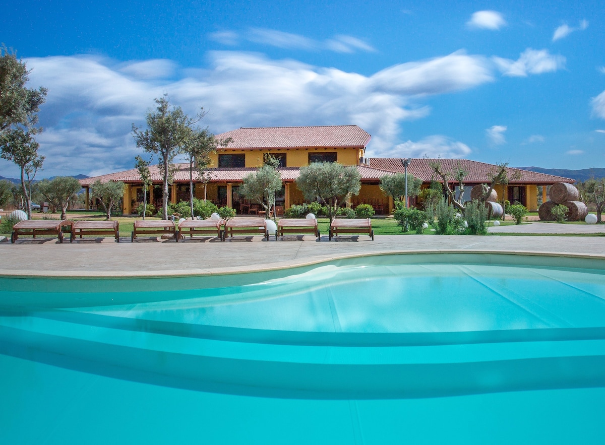 An outdoor pool is visible in the foreground, with clear turquoise water reflecting the sky. In the background, a two-story yellow building is surrounded by olive trees and manicured gardens. Benches are positioned along the poolside, inviting relaxation.