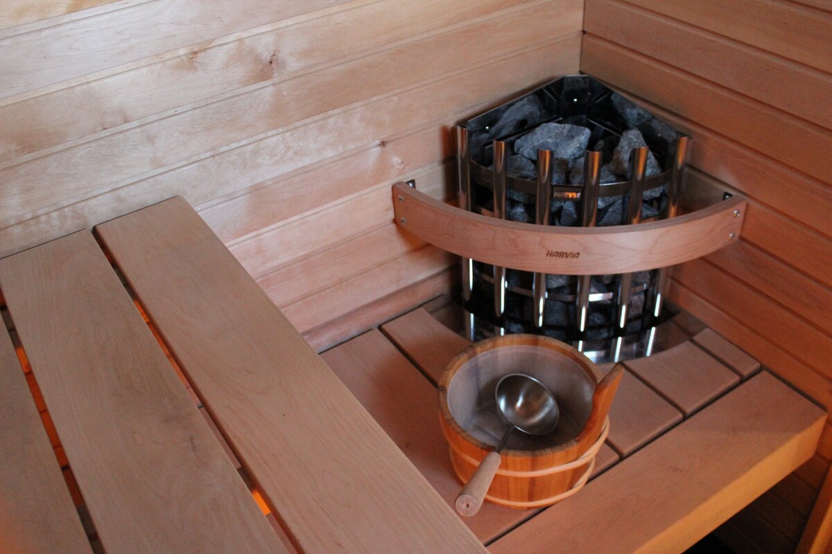 A sauna area is shown, featuring warm wooden benches arranged in a corner. A stone heater is visible against the wall, surrounded by a wooden surround. A wooden bucket and ladle are placed nearby, inviting relaxation and wellness.