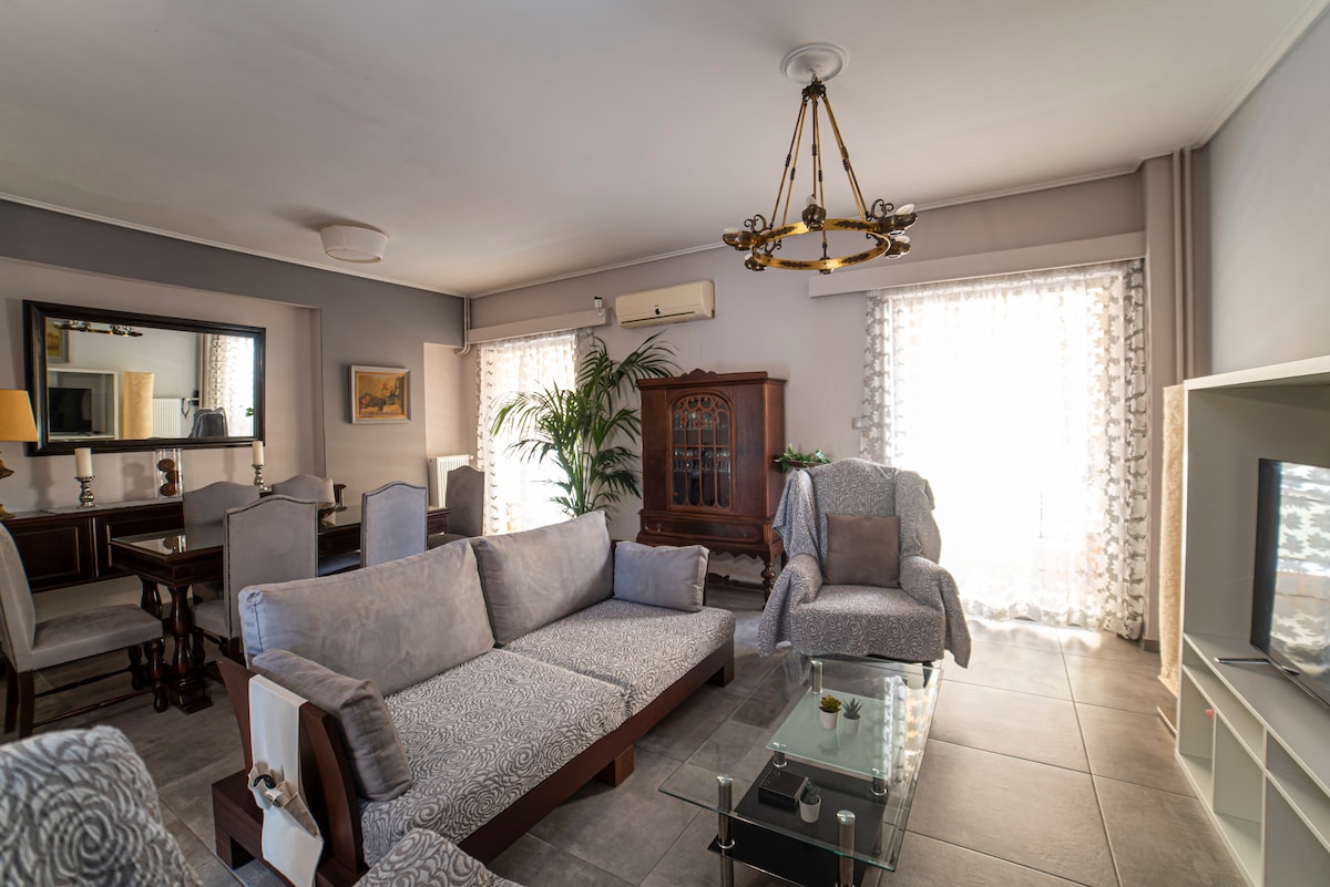 A spacious living area features a light-colored sofa and a patterned armchair, complemented by a glass coffee table. Natural light enters through sheer curtains, highlighting a decorative mirror and a wooden cabinet. A dining table with chairs is visible in the background.