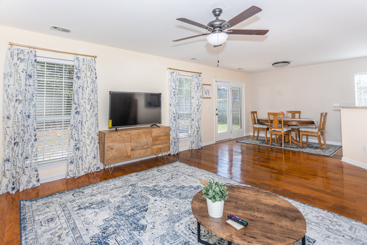 The living room features hardwood floors and large windows that provide natural light. A comfortable seating area is arranged around a coffee table, and a 55" smart TV is mounted on the wall. A dining area is visible in the background, complemented by decorative curtains.