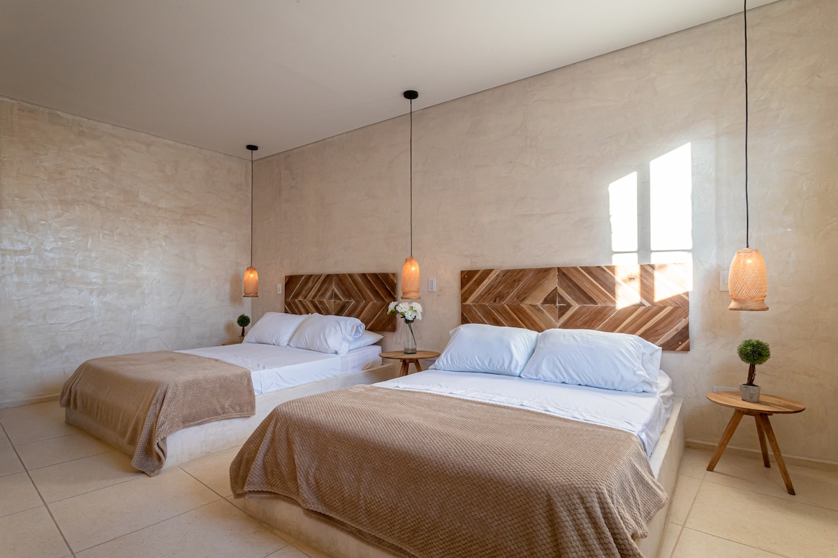A spacious sleeping area is furnished with two comfortable beds featuring light-colored linens. Wooden headboards add a natural touch, while bedside tables hold small decorative plants. Soft pendant lights hang above each bed, and natural light enters through a window, enhancing the serene ambiance.