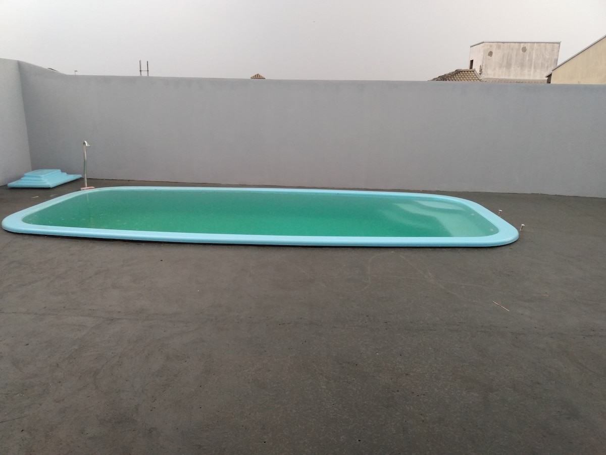 A spacious swimming pool is framed by a light blue border, reflecting a calm green hue. The surrounding area is paved, providing ample space for relaxation. The wall in the background is painted a soft gray, offering a neutral backdrop to the pool.