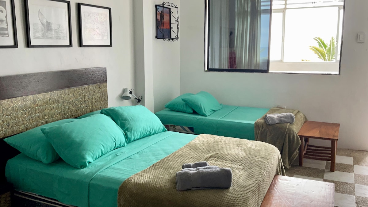 A cozy sleeping area features two spacious beds adorned with soft turquoise bedding. A wooden side table holds neatly arranged towels. Natural light floods the room through a large window, providing a view of greenery outside and enhancing the inviting atmosphere.