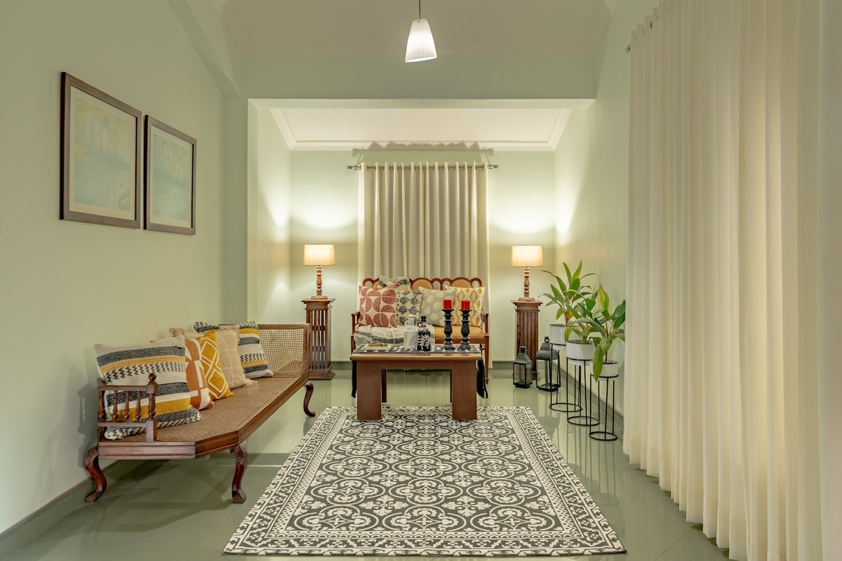 A spacious living room is illuminated softly by table lamps, featuring a comfortable sofa adorned with patterned cushions. Light curtains frame large windows, and a decorative rug anchors the seating area. Potted plants bring a touch of greenery to the serene ambiance.