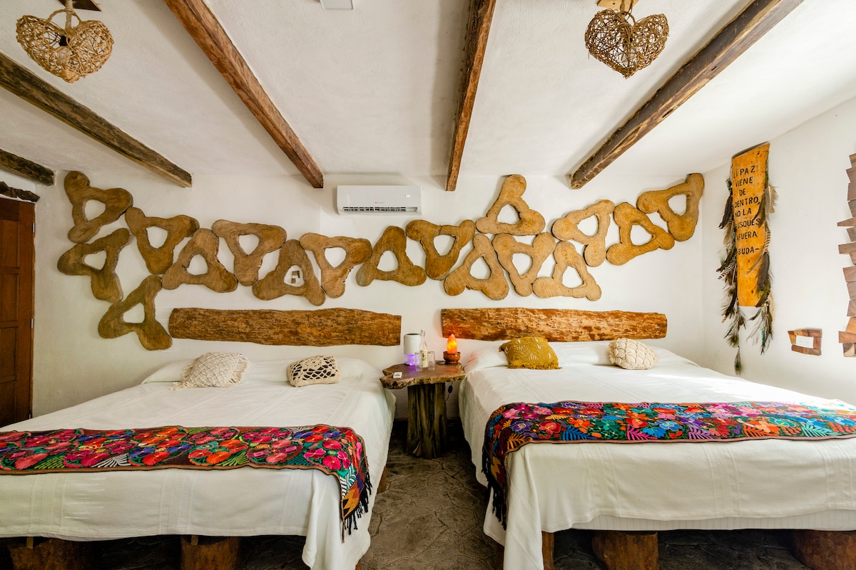 A cozy room features two spacious beds with white linens, accented by a vibrant embroidered blanket. A wooden artistic wall decor enhances the rustic charm. Natural light filters in through a nearby window, and unique light fixtures made of woven material hang from the ceiling.