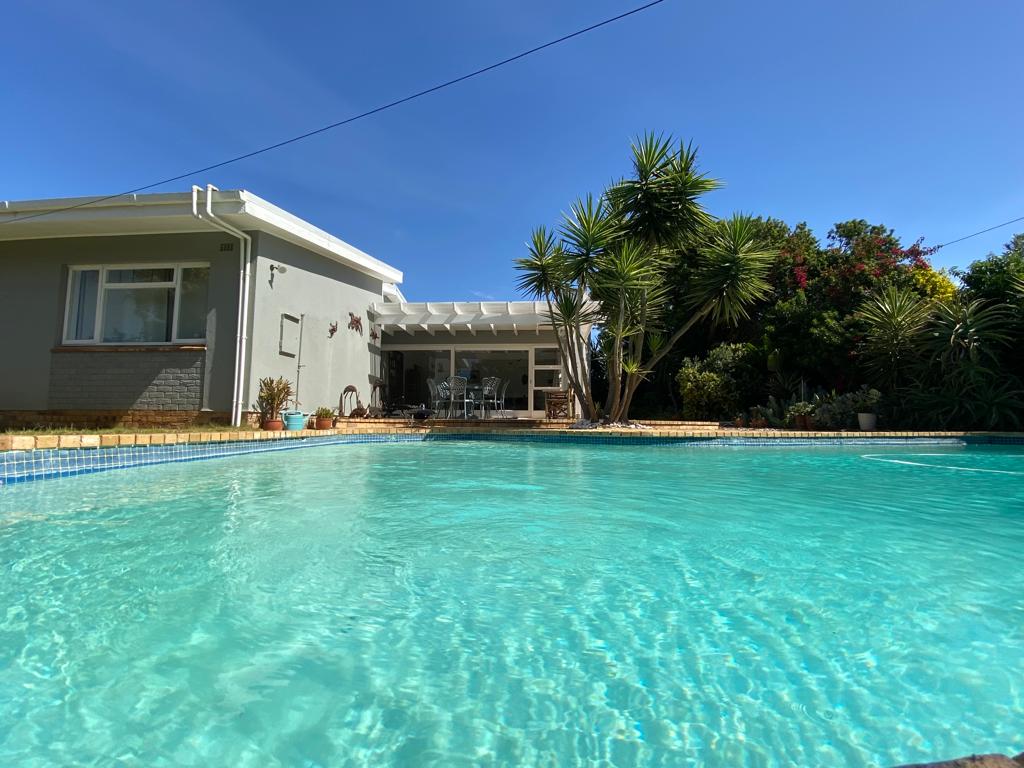 A clear and inviting pool reflects the bright blue sky, surrounded by lush tropical plants and trees. The house is visible in the background, showcasing a spacious patio area for outdoor dining and relaxation.
