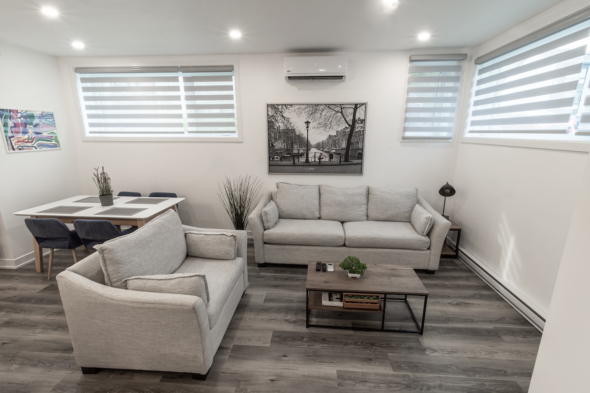 A modern living area is presented with two light gray sofas facing a low coffee table. A dining table with four chairs is positioned to one side, while a piece of artwork adorns the wall. Natural light filters through the striped window coverings.