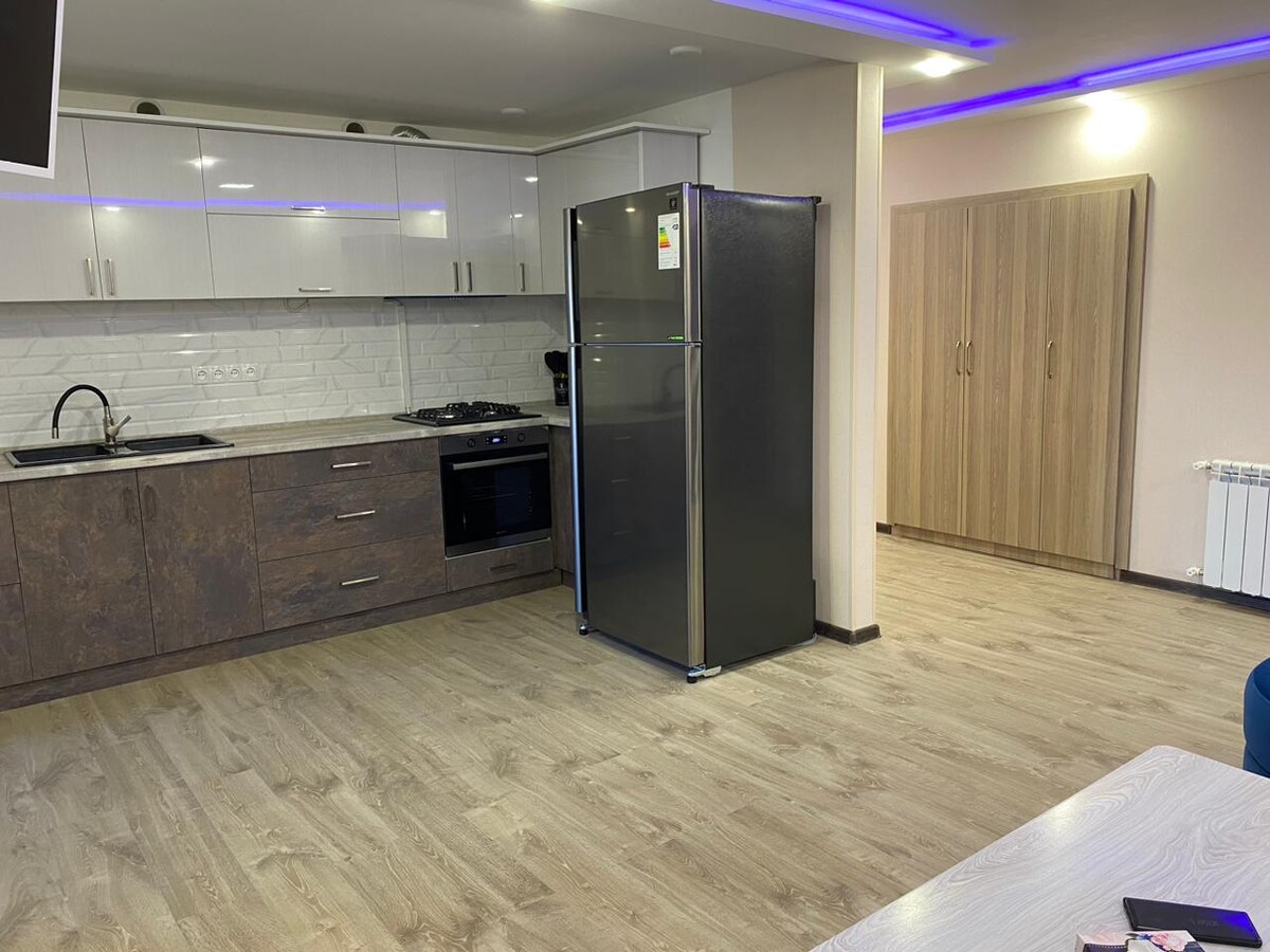 The kitchen area features modern cabinetry in a light tone, complemented by a contrasting dark countertop. A stainless steel refrigerator stands alongside a built-in oven and cooktop. The open space is illuminated by subtle ceiling lights, showcasing neutral-toned flooring.