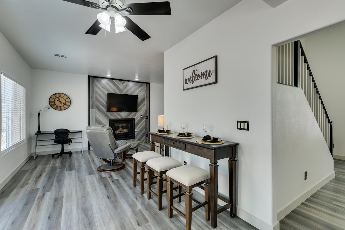 A cozy living space features a comfortable seating area with a recliner and a fireplace. A rustic table with four stools is set for dining. A wall clock adds charm, while large windows allow natural light to illuminate the room.