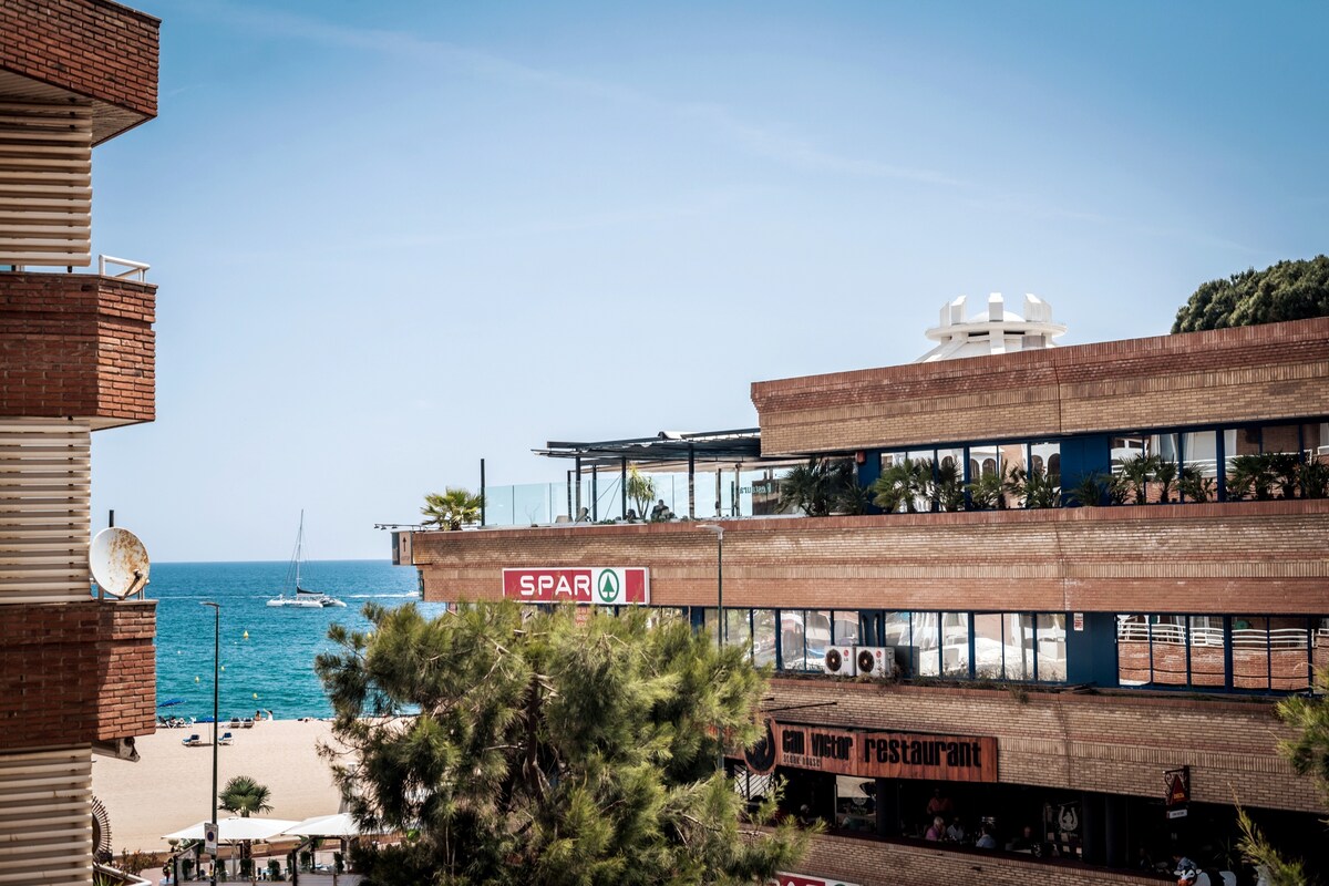 A view of the coastline shows the nearby beach and distant sailing boat beneath a clear blue sky. Beachfront establishments are visible, including a restaurant and a grocery store. Lush greenery provides a contrast to the urban setting, enhancing the tranquil atmosphere.