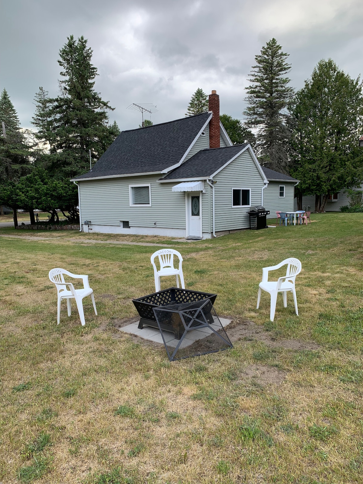 A gathering area is arranged with four white plastic chairs positioned around a black fire pit, situated on a flat green lawn. The backyard features a simple grey house with a sloped roof and a small window. Trees are visible in the background.