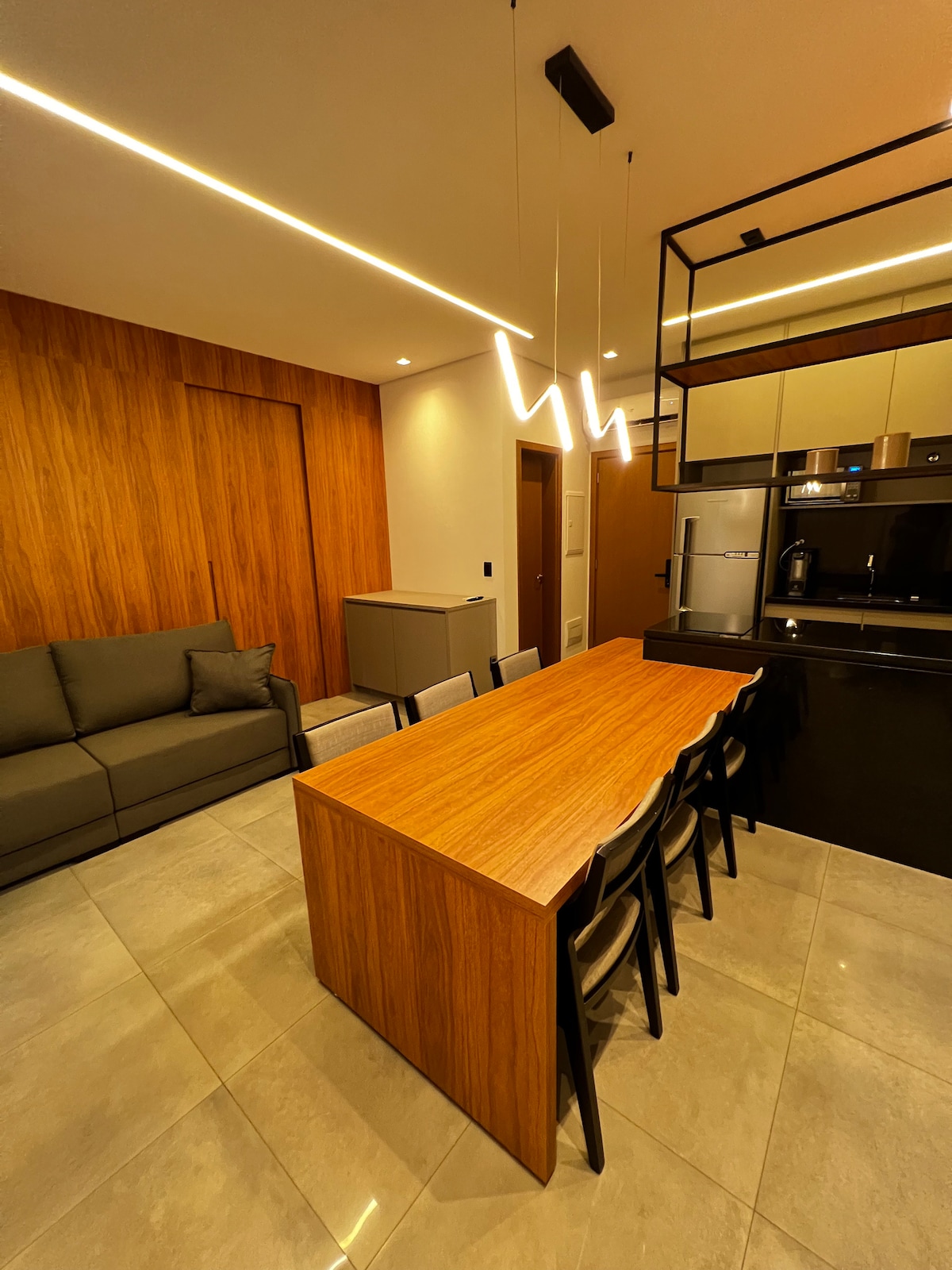 A modern dining area is highlighted by a long wooden table surrounded by black chairs. The space features a sleek kitchen with dark cabinetry and appliances, while a comfortable sofa is positioned against a wooden accent wall, complemented by ample lighting.