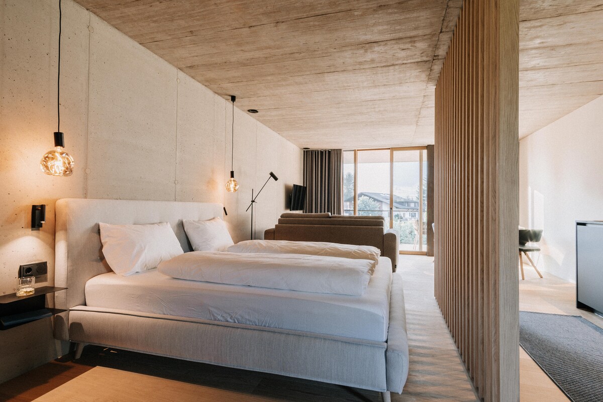 An open living and sleeping area features a spacious bed, complemented by modern pendant lighting. A partial wooden divider delineates the space, while large windows allow natural light to flood in, revealing a view of the surrounding landscape.