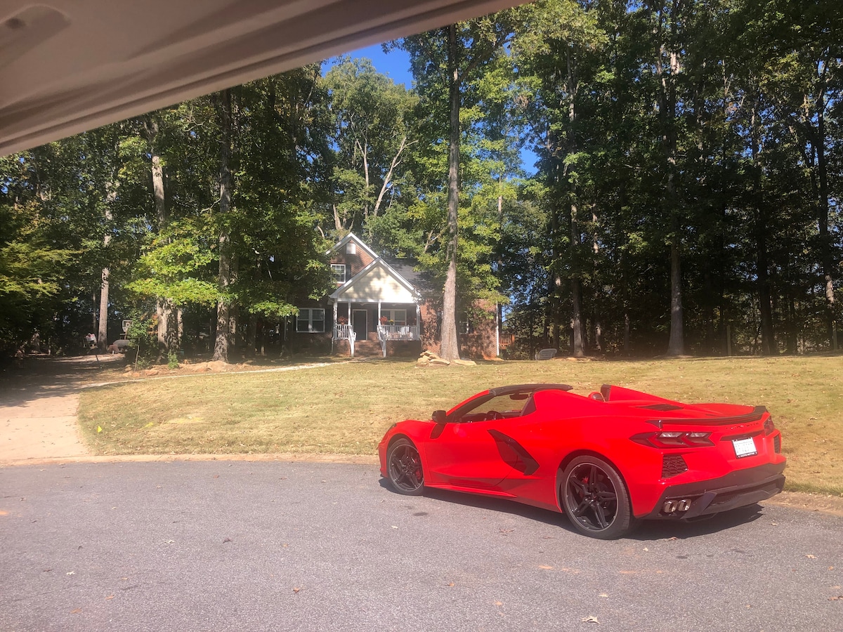 A striking red sports car is parked in the foreground, with a spacious lawn and trees in the background. The lodge-style house, with multiple windows, is visible among the trees, providing a serene setting.