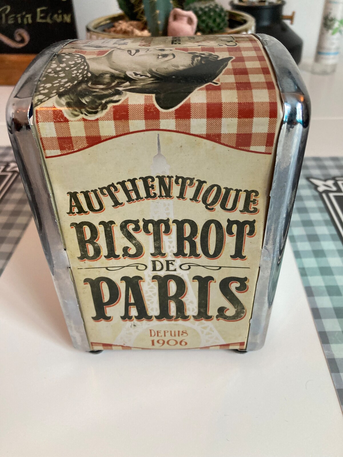 An antique-style metal container is displayed on a patterned tablecloth. The container features a vintage design with a red-and-white checked border and illustrations depicting a Parisian theme. Text indicates it is associated with the 'Authentique Bistrot de Paris' and has a date of 'Depuis 1906'.