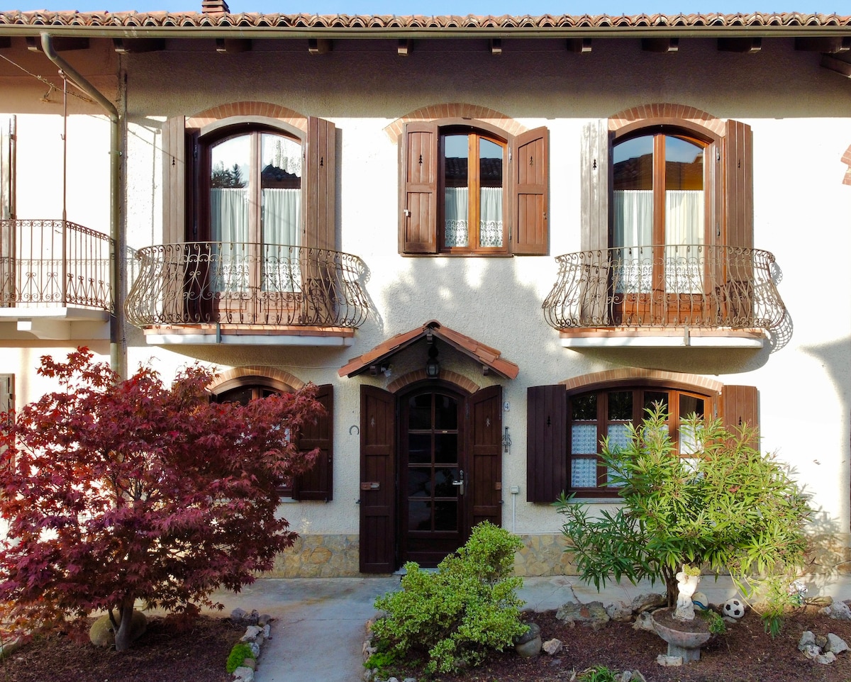 The exterior of the rustic house showcases traditional elements, including intricate wrought iron balconies and wooden shutters. A well-maintained garden with shrubs and small trees adds a touch of greenery, while the inviting entrance features a decorative wooden door under a small archway.