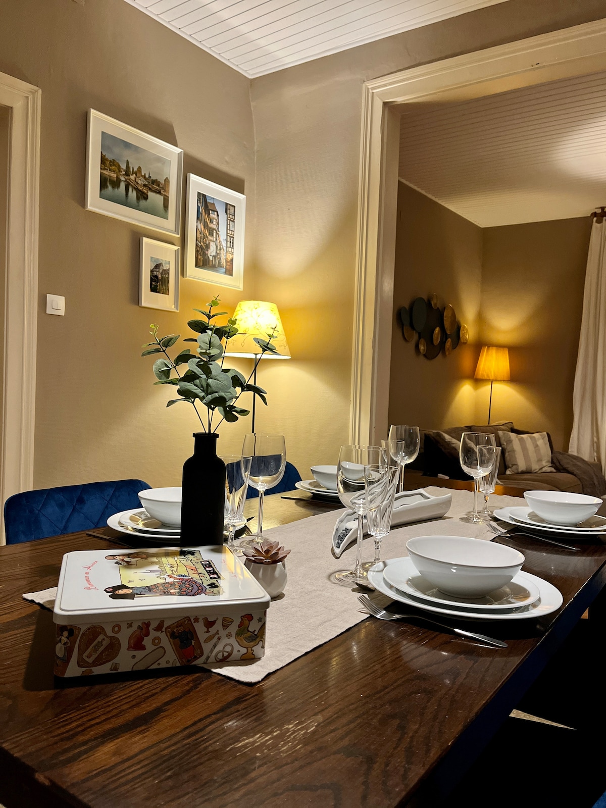 A dining table is set with white plates and glassware, accompanied by a decorative vase with greenery. Soft lighting from a nearby lamp provides a warm ambiance, while framed art is displayed on the walls, enhancing the cozy atmosphere of the dining area.