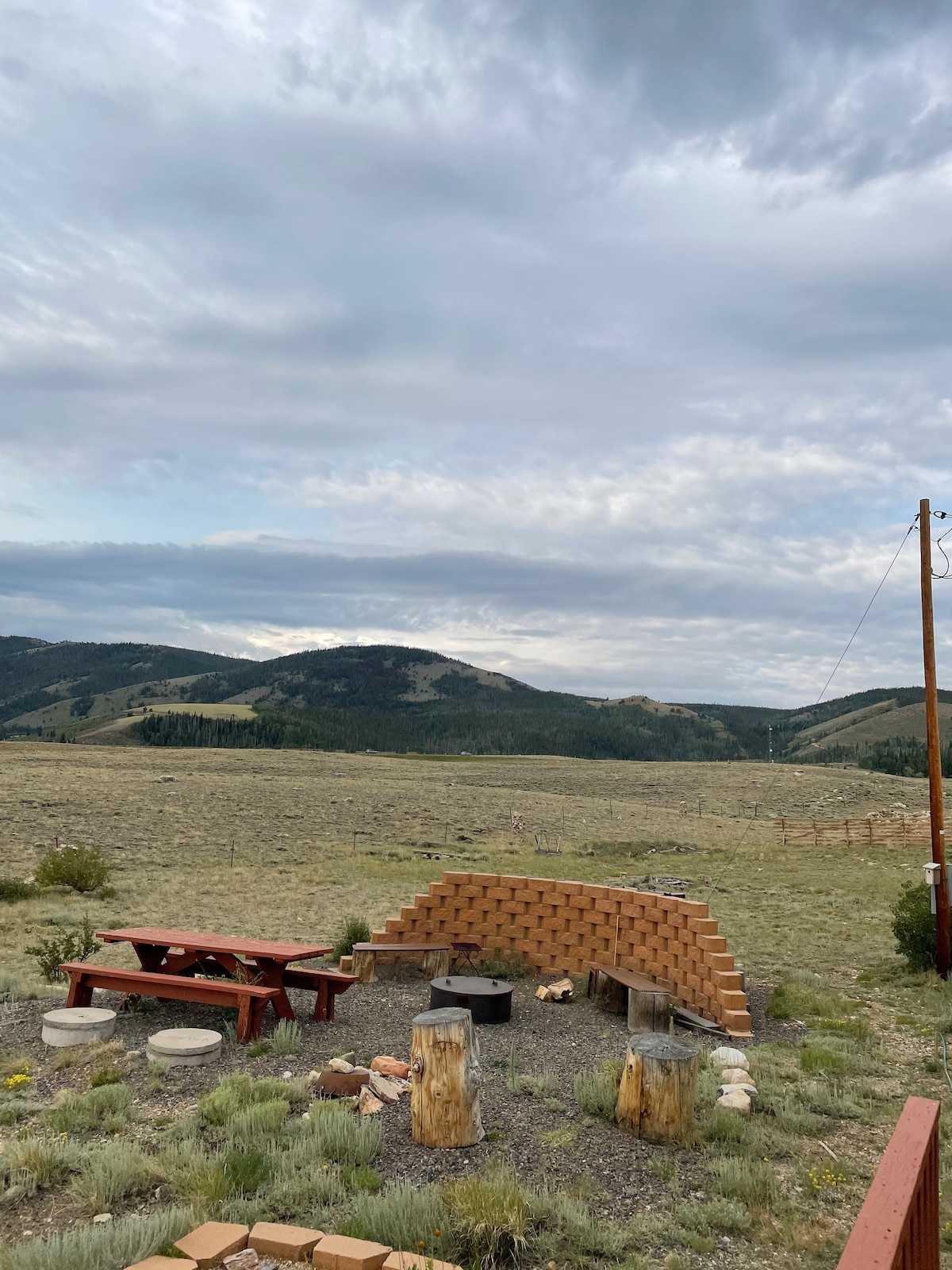 An outdoor fire pit area is surrounded by natural landscaping, featuring a brick fire ring and log seating. The scenic view includes rolling hills under a clouded sky, providing a vast open space ideal for enjoying evenings around the fire.