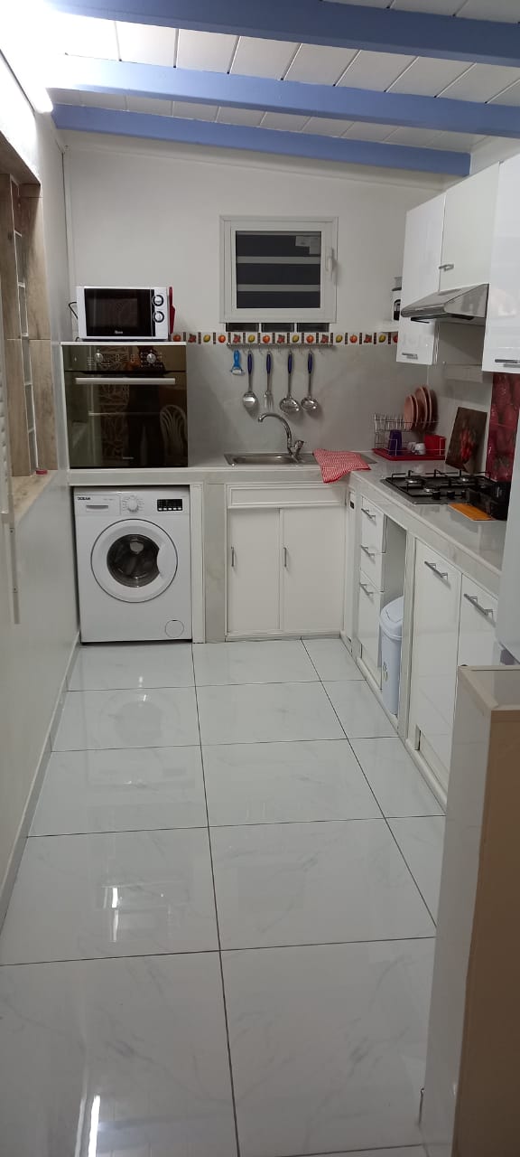 A modern kitchen features white cabinetry and sleek countertops. A washing machine is positioned next to the sink, with cooking utensils neatly arranged on the wall. Natural light enters through a window, enhancing the clean and organized appearance of the space.