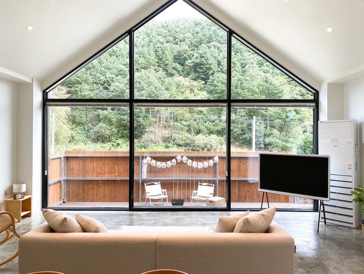 A spacious living area is highlighted by a large triangular window, offering views of surrounding greenery. A modern sofa faces a flat-screen TV, with light-colored decor creating a bright atmosphere. Two outdoor chairs are visible on the balcony, enhancing the inviting space.