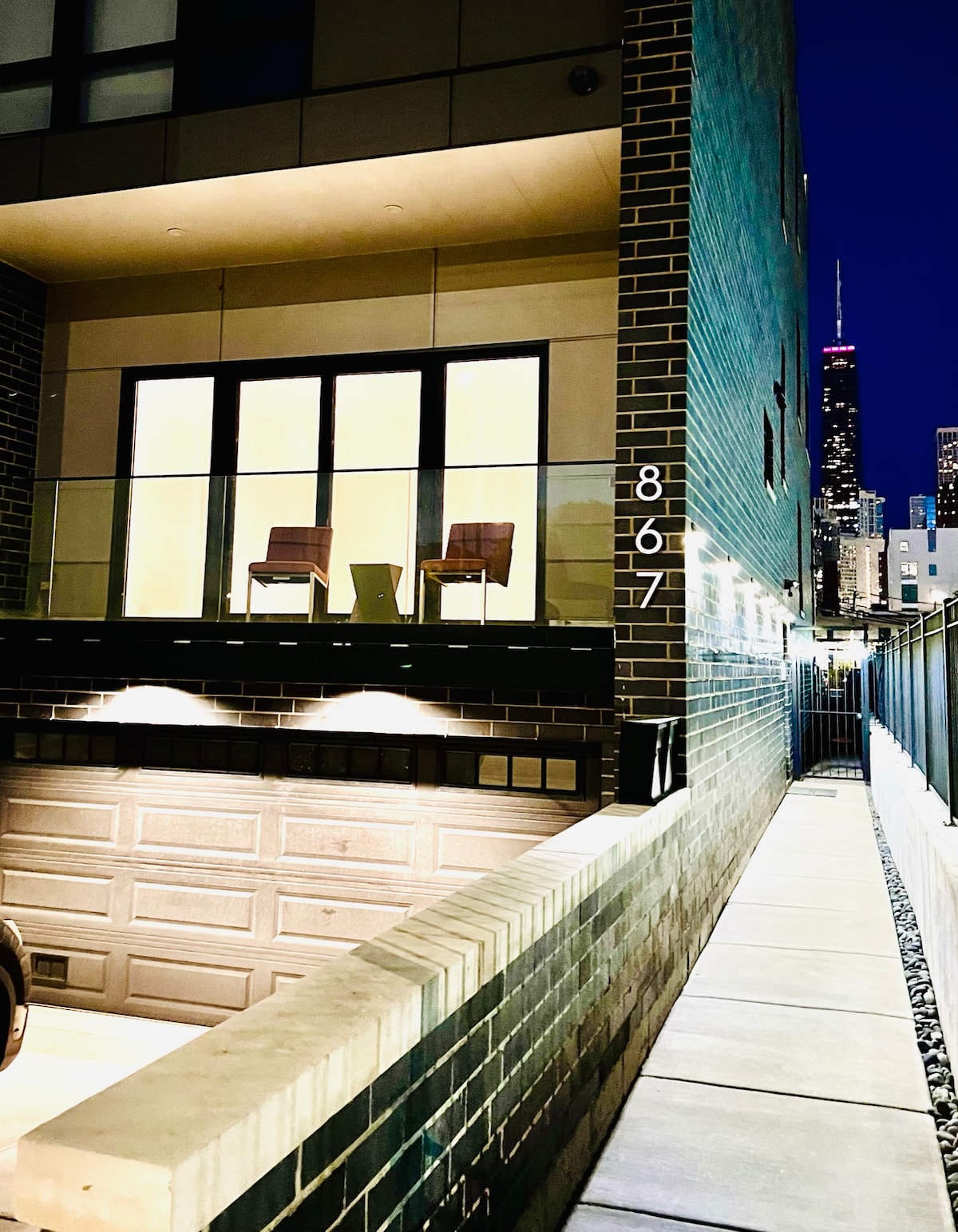 An exterior view of the modern apartment building is showcased at night. Bright lights illuminate a balcony with two chairs overlooking a walkway. The Chicago skyline, including the Willis Tower, is visible in the background, enhancing the urban ambiance.