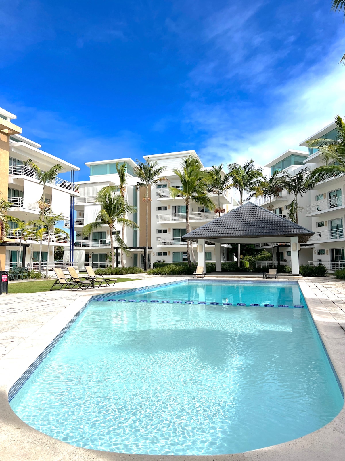 A clear pool sparkles under a bright blue sky, surrounded by lush palm trees and modern buildings. Sun loungers are positioned around the pool area, providing a relaxing space for guests. An inviting gazebo is situated near the pool, offering shaded seating.