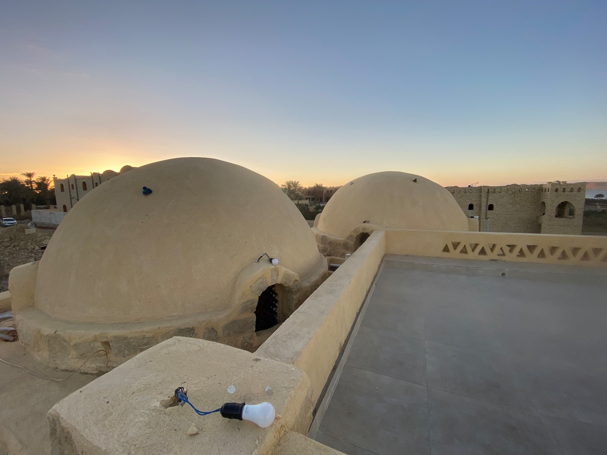Two curved domes are visible against a serene sunset, showcasing the unique architectural style of the building. A flat rooftop space offers an expansive view, with warm hues of the evening sky in the background. A light bulb is placed on the ground, indicating outdoor lighting.