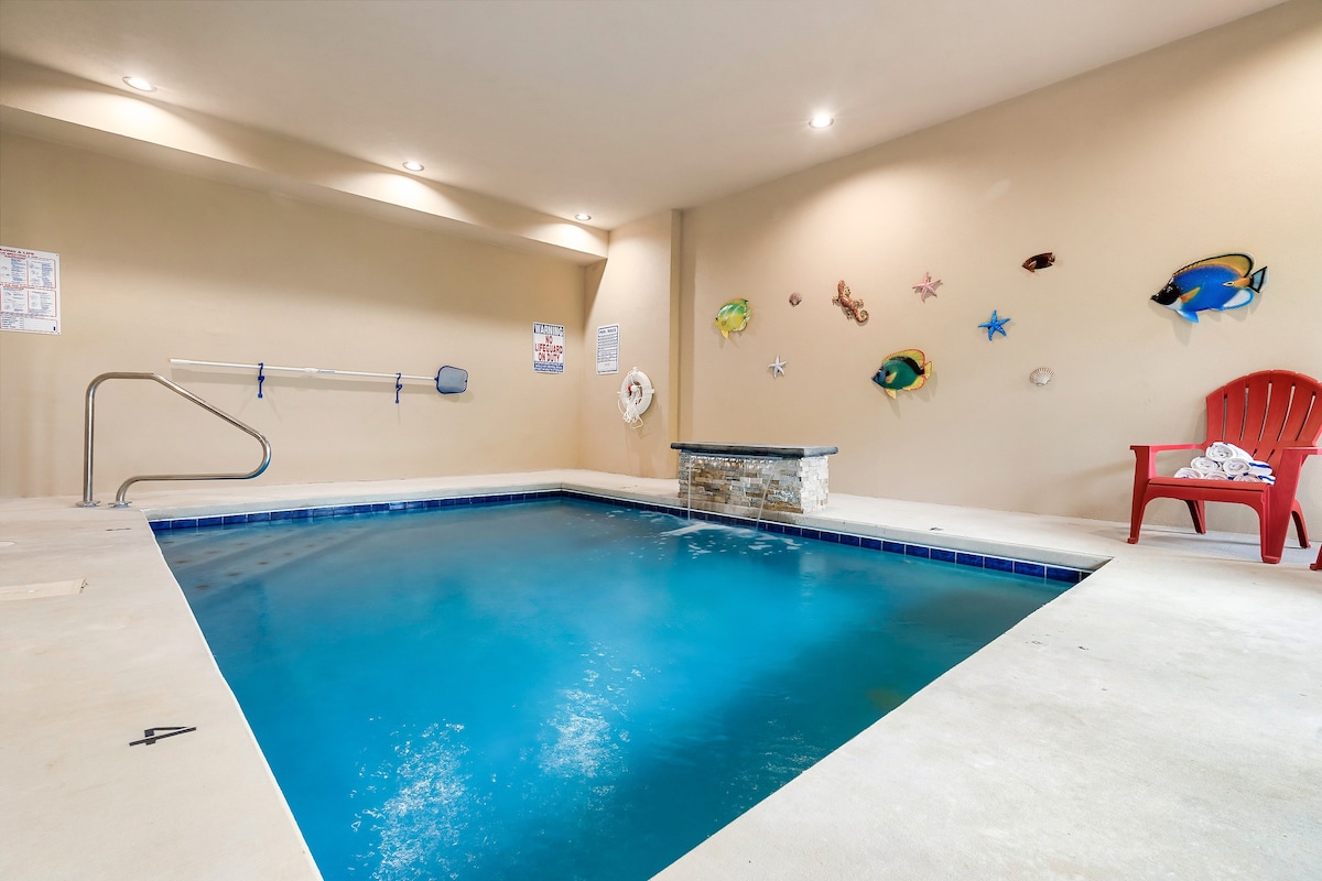 An indoor pool is featured in a well-lit area, with a deep blue water surface and an accessible entry step. Colorful fish decorations adorn the walls, while a red chair sits adjacent to the pool. The pool is surrounded by a smooth concrete floor, providing ample space.
