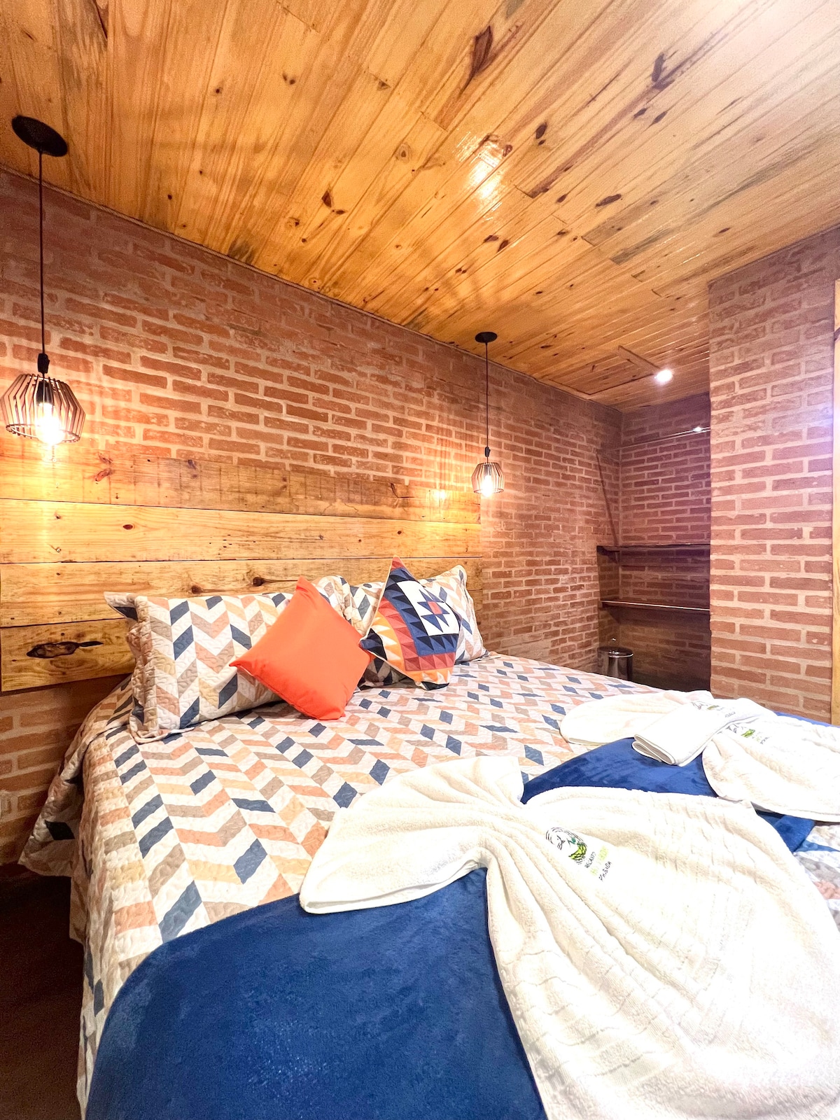 A rustic bedroom features warm wooden walls complemented by exposed brick and a spacious bed adorned with patterned bedding. Decorative pillows in vibrant hues are arranged on the bed. Soft lighting is provided by pendant lamps, enhancing the cozy ambiance.