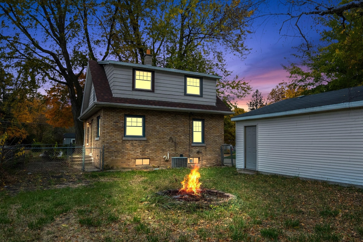 The exterior of the cozy cottage is viewed at twilight, with warm light glowing from the windows. A fire pit occupies the fenced backyard, surrounded by grass. The structure features a two-story design, with a shed visible to the right.