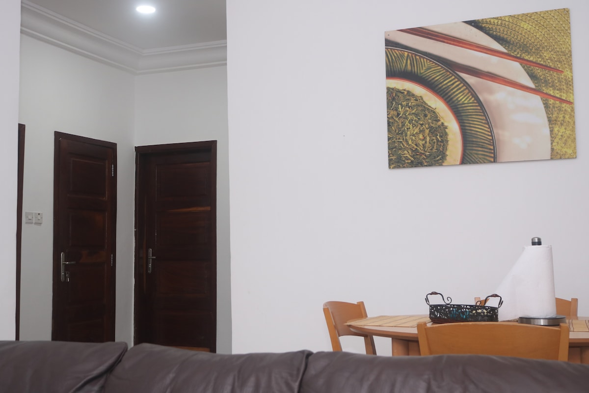 A cozy living space is shown, featuring a wooden dining table with four chairs next to a decorative wall art piece. Two doors lead to other rooms, and soft lighting illuminates the area, highlighting the modern furnishings.