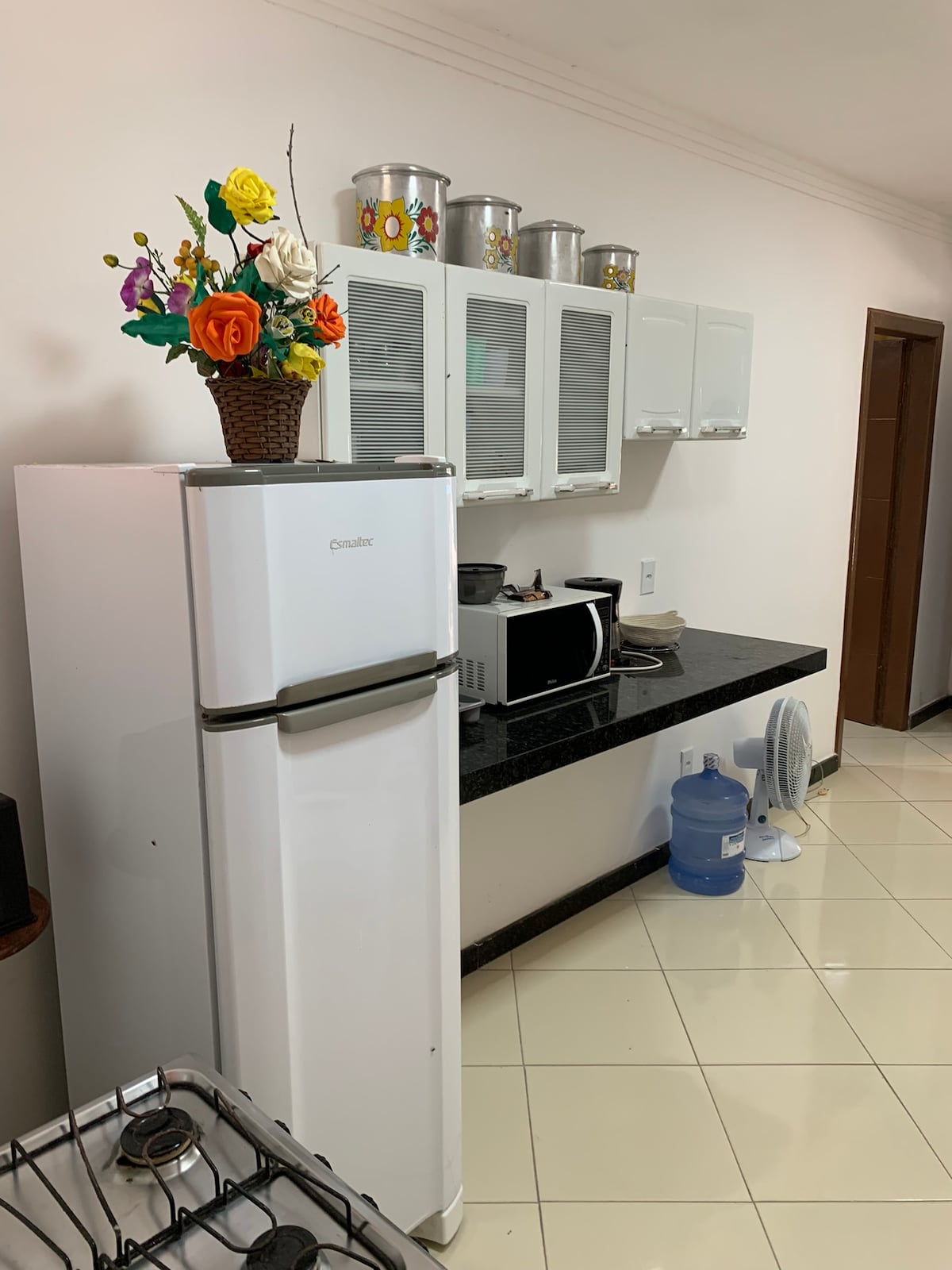 A functional kitchen space is featured, with a white refrigerator and a countertop microwave. Above the counter, cabinets with glass panels showcase decorative items. A water dispenser and a fan are positioned beside the fridge, while a gas stove is located in the foreground.