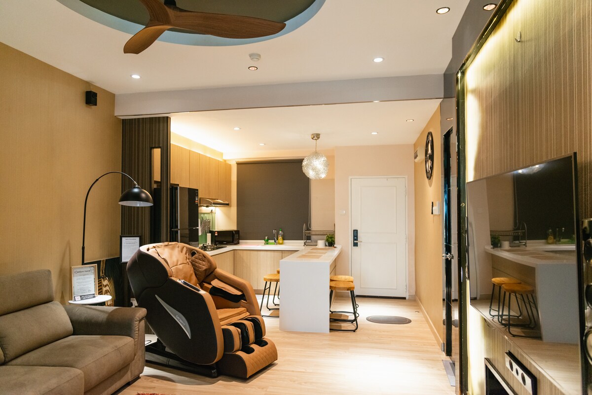 An open living area features a massage chair and a comfortable sofa. A modern kitchen with wooden cabinetry is visible in the background, accompanied by bar stools at the island. Soft lighting creates a relaxed atmosphere, complemented by a large screen TV against one wall.