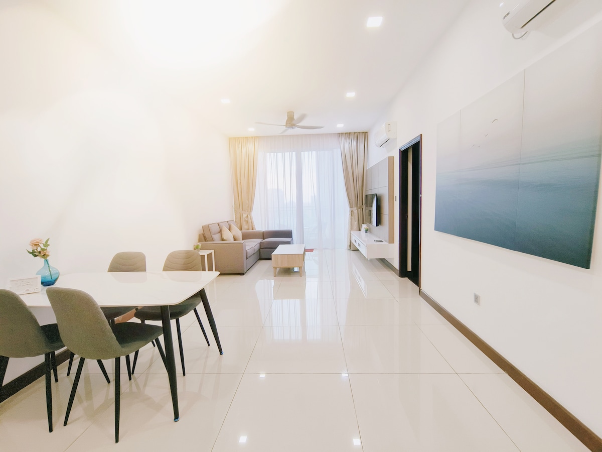 A spacious living area is highlighted by large windows dressed in soft curtains, allowing ample natural light. A modern sofa arrangement is positioned alongside a sleek entertainment unit with a TV. A dining table with four chairs is visible, complementing the glossy tile flooring.