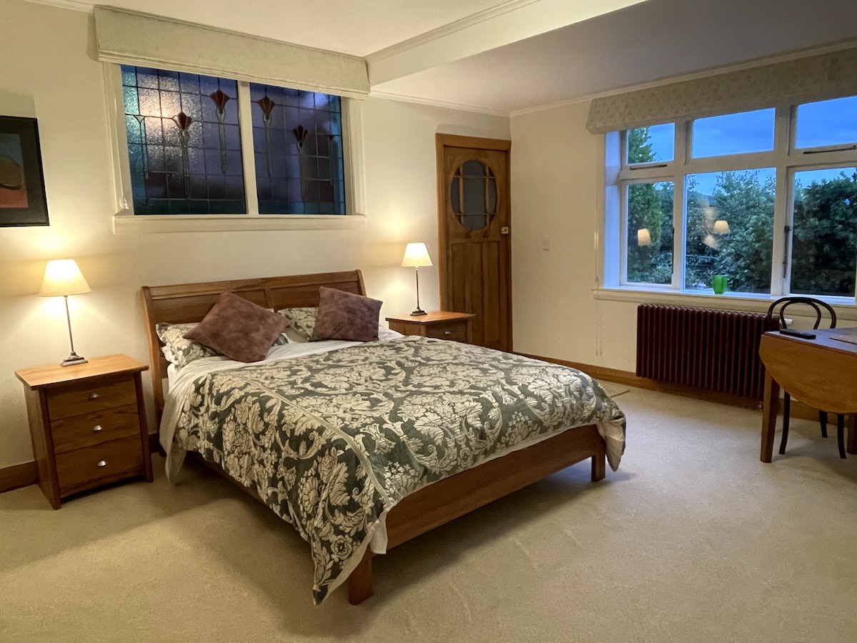 A spacious bedroom features a large wooden bed adorned with a patterned blanket and two decorative pillows. Two bedside tables with lamps flank the bed. A cozy seating area includes a small table and chair. Large windows allow natural light to illuminate the room.