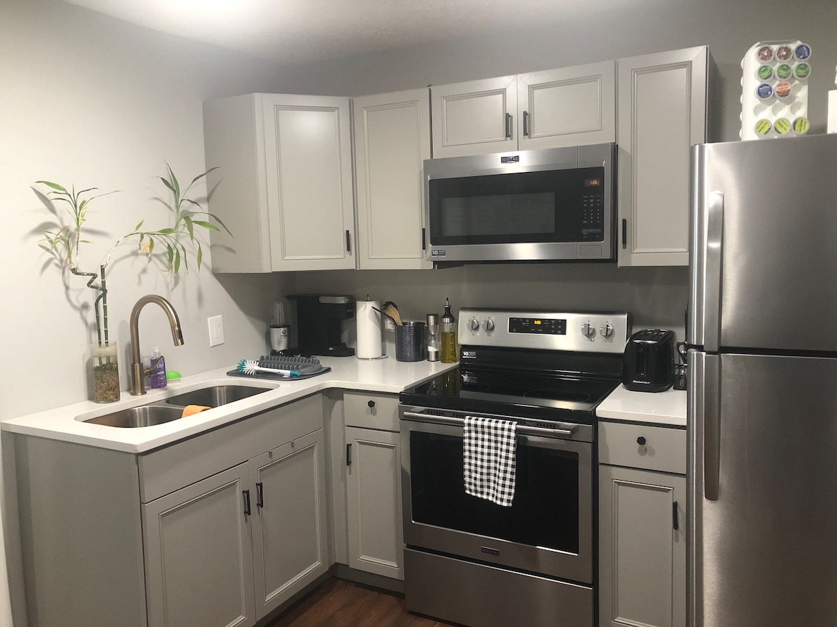 A modern kitchen features gray cabinetry and stainless-steel appliances, including an oven, microwave, and refrigerator. A sleek sink is complemented by a stylish faucet. Natural light is enhanced by neutral wall tones, while a small plant adds a touch of greenery to the space.